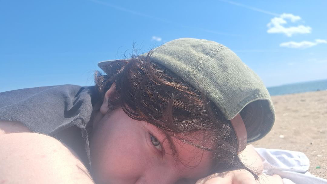 a picture of rhys, a boy with fair skin and brown hair tucked under a green baseball cap, laying on a beach.